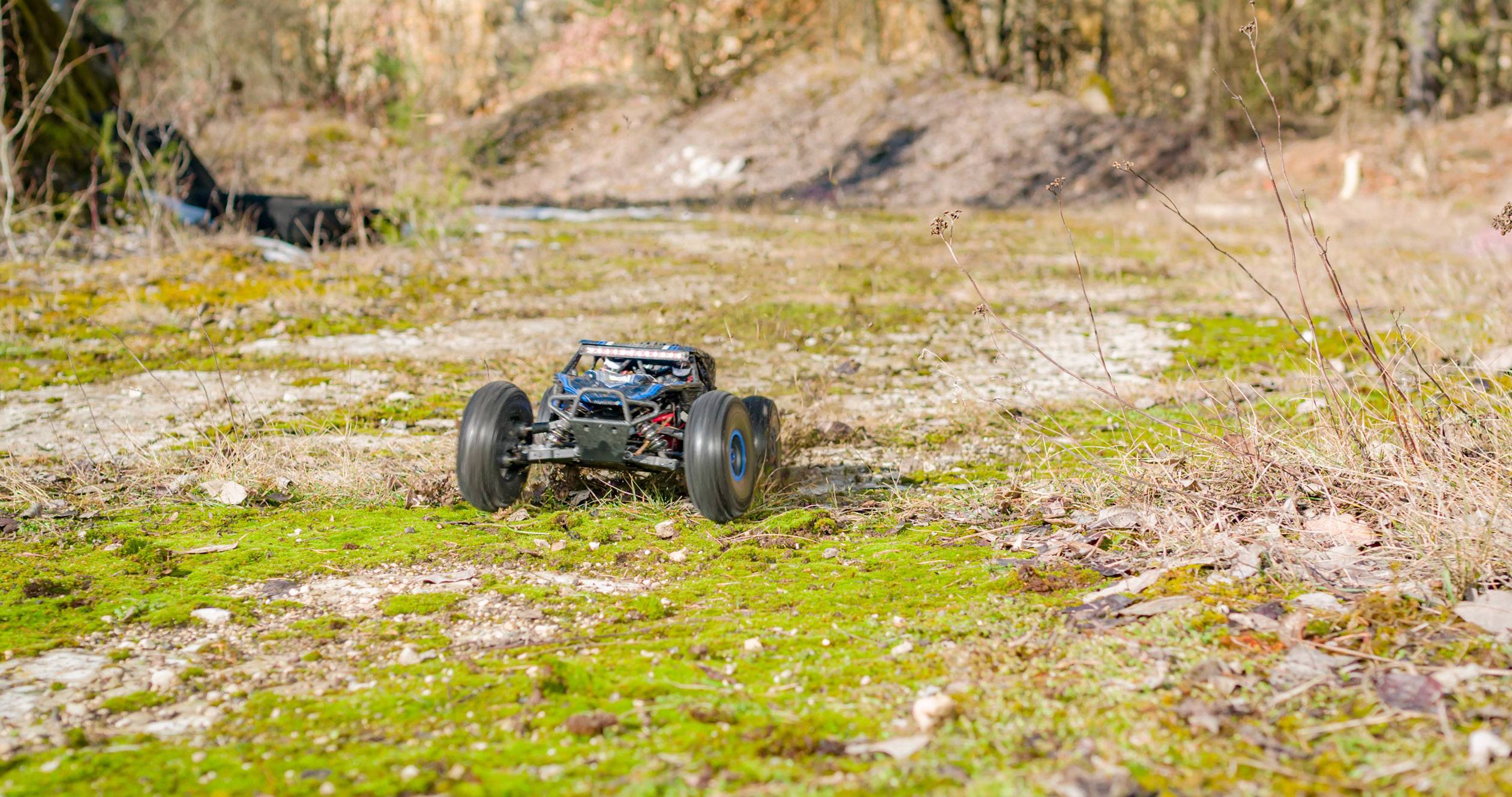 Een op afstand bestuurd auto rijdt snel over een met mos begroeid, stenig pad in een natuurlijke omgeving met bomen op de achtergrond.