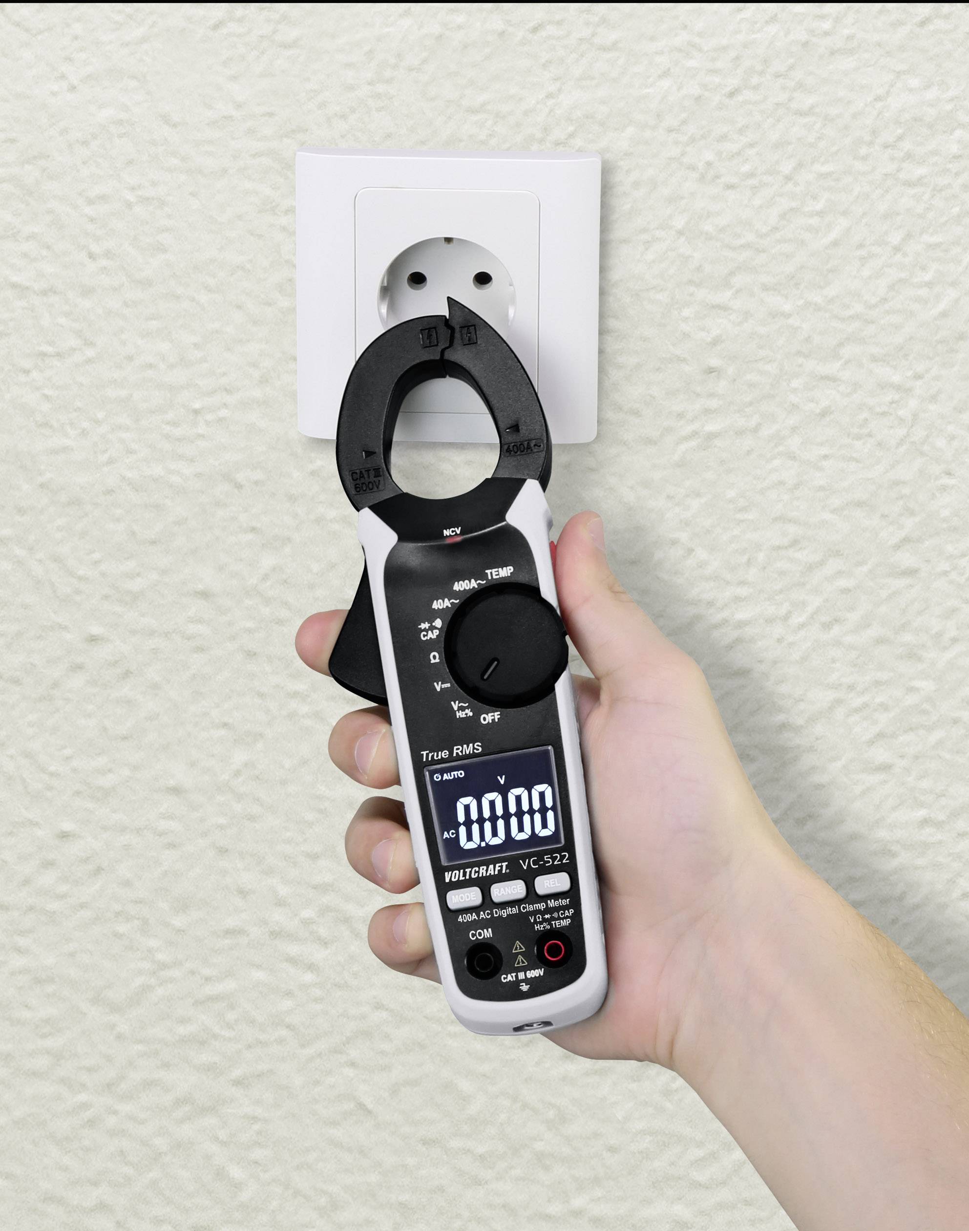 Een hand houdt een stroommeter voor een witte wandcontactdoos. Het apparaat toont '0000' op het digitale display.