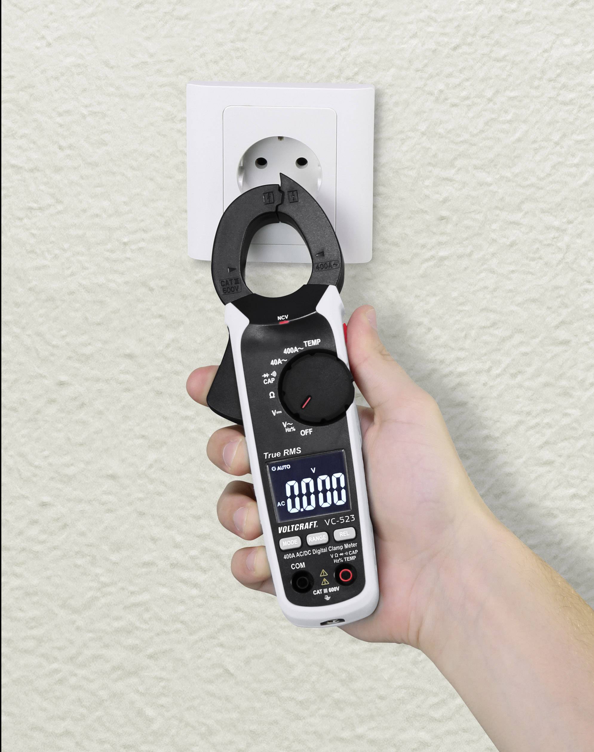 Een hand houdt een multimeter voor een stopcontact. Het display toont '0.00'. Het apparaat meet elektrische stromen en spanning.