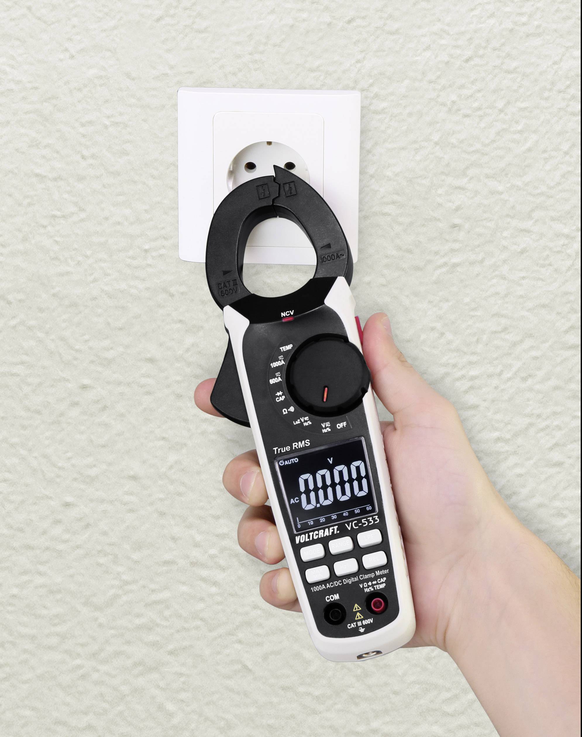 Een hand houdt een multimeter voor een stopcontact, waarbij het display 0,00 aangeeft. Het apparaat wordt mogelijk gecontroleerd op spanning.