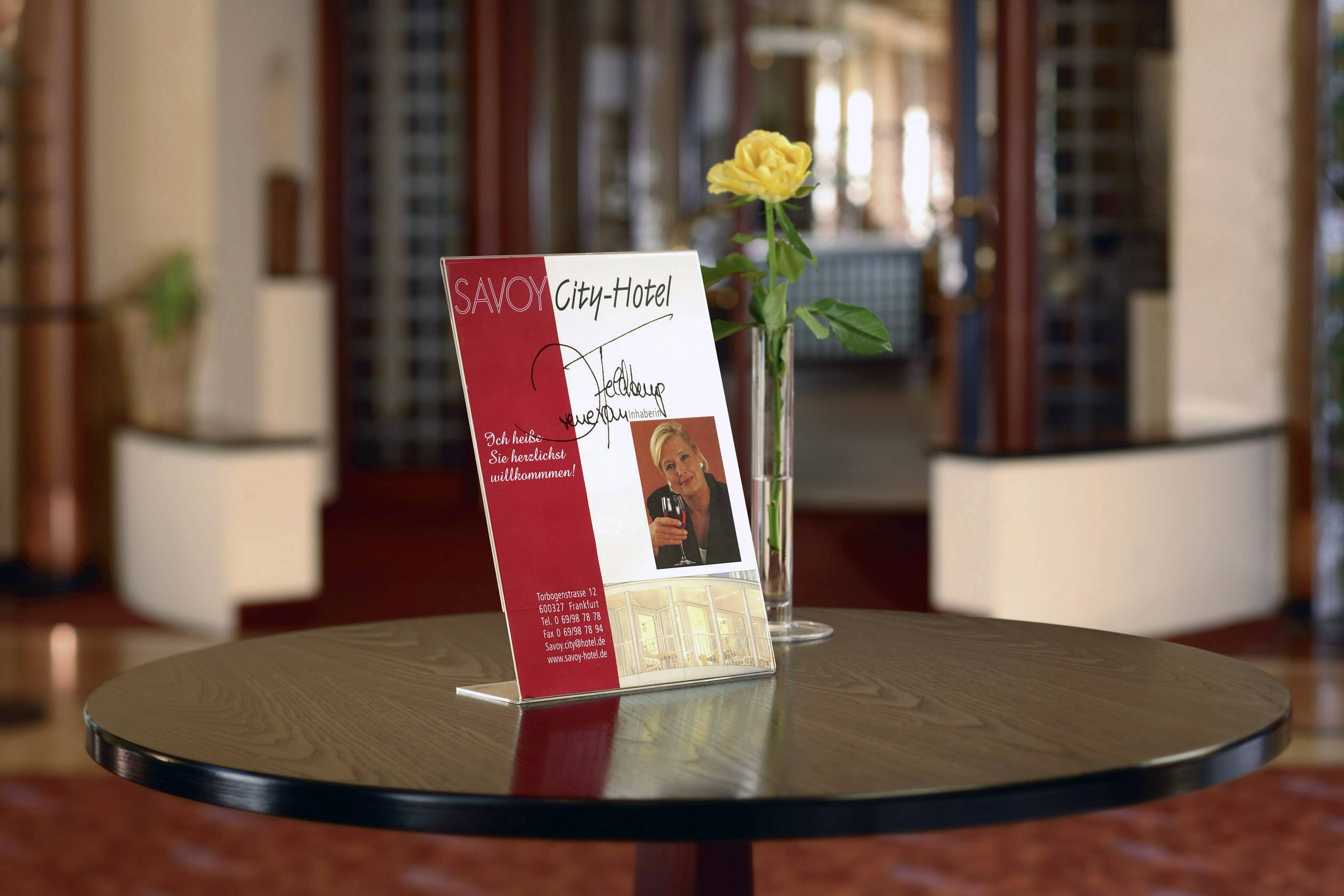 Hotelbrochure op tafel presenteert aanbiedingen en services van het Savoy City Hotel. Naast de brochure staat een gele roos als decoratie. Op de achtergrond is het hotelinterieur zichtbaar.