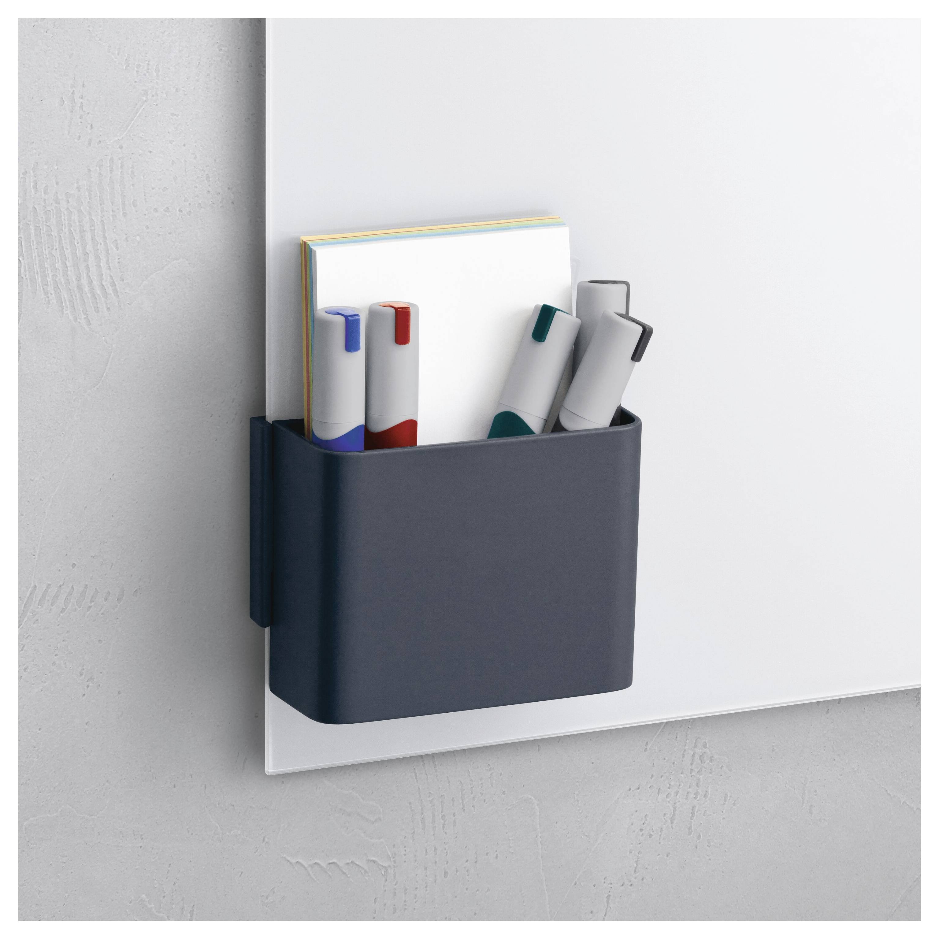 Aan de muur bevestigde donkergrijs organizer met notitieboekjes en markeerstiften op een wit paneel tegen een lichtgrijze wand.
