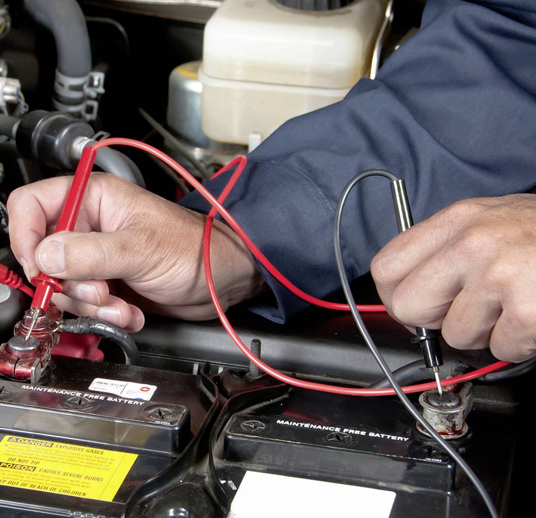 Een persoon meet de spanning van een autobatterij met twee meetpennen, rood en zwart, aan de batterijpolen in de motorruimte.