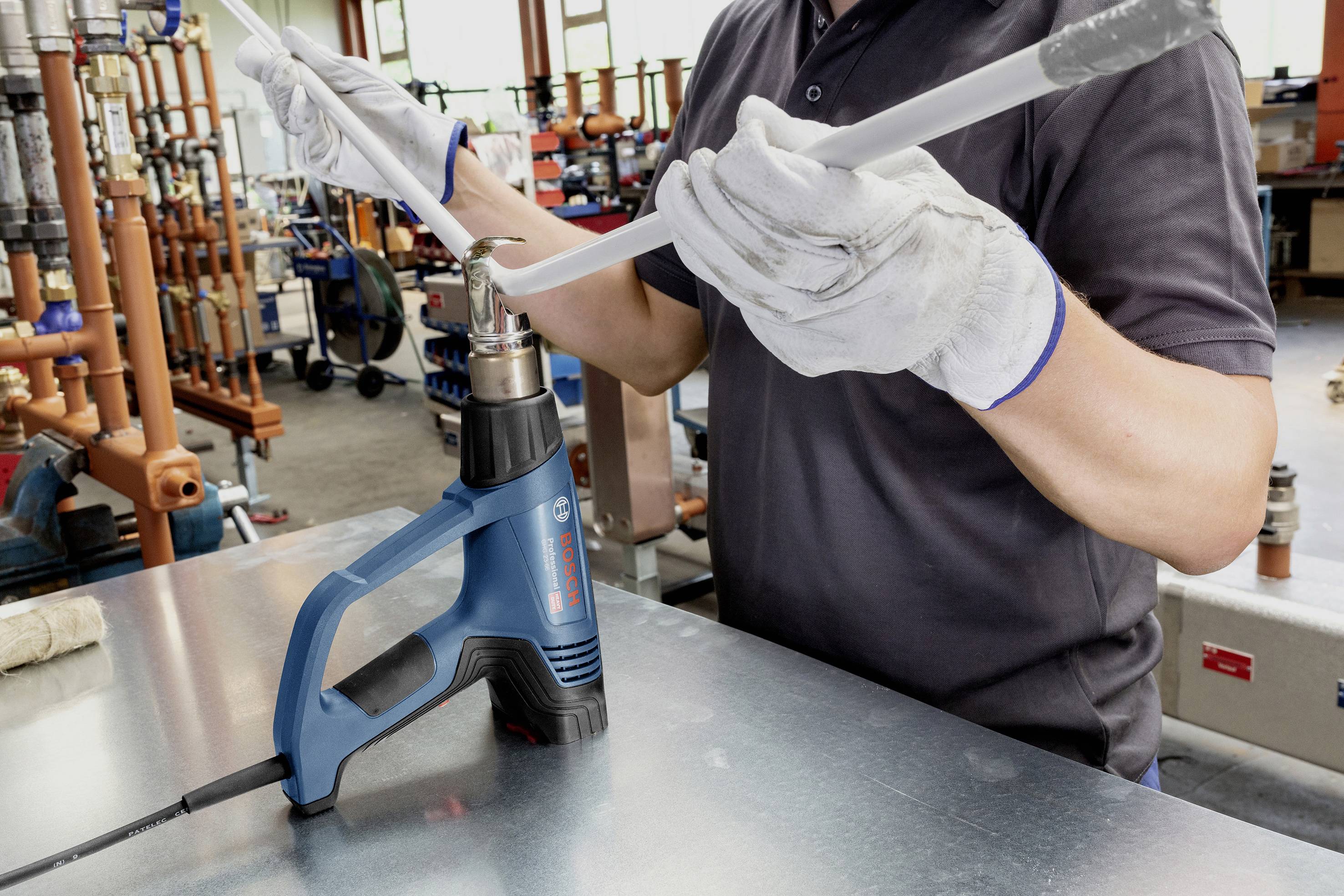 Een persoon met handschoenen verhit een kunststofbuis met een heteluchtpistool in een werkplaats vol sanitaire onderdelen.