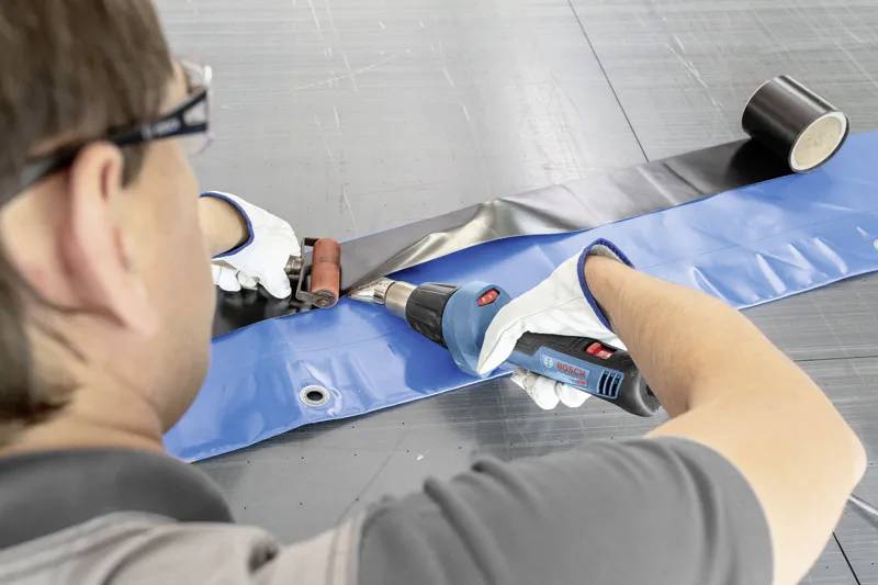 Een persoon gebruikt een heteluchtpistool en een rol om een naad in dakbedekkingsmateriaal te lassen, terwijl hij veiligheidshandschoenen en een veiligheidsbril draagt op een grijze ondergrond.