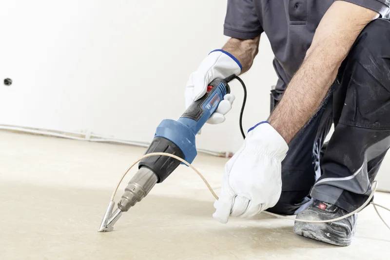 Een persoon gebruikt een heteluchtpistool op een kabel die op de vloer ligt, terwijl hij beschermende handschoenen en werkkleding draagt, gefocust op een constructietaak.