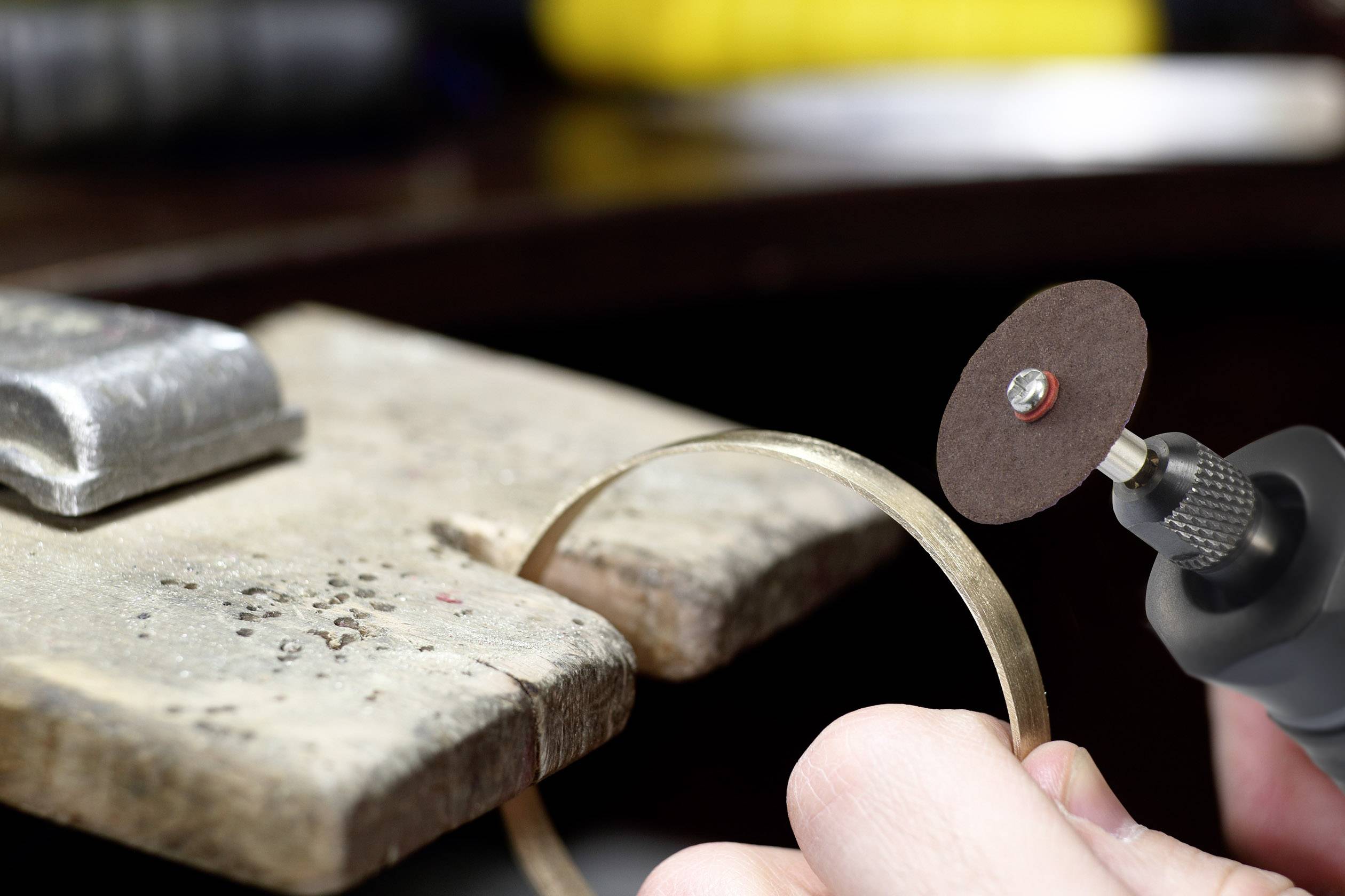 Een hand houdt een roterend slijpgereedschap vast om een gouden metalen ring te bewerken, die op een houten blok ligt.