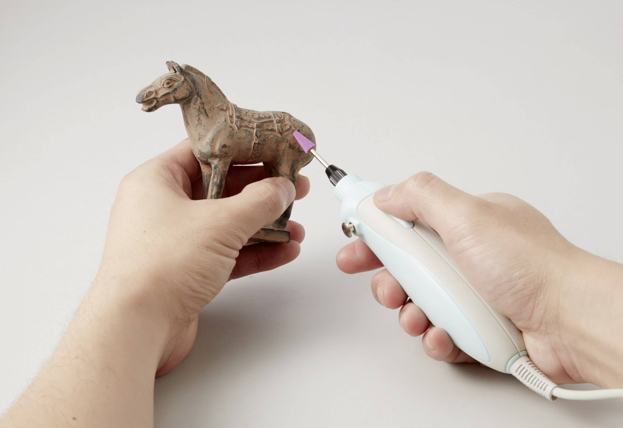 Een persoon houdt een paardensculptuur in de ene hand en gebruikt een elektrisch slijpgereedschap in de andere hand om het beeld te bewerken.
