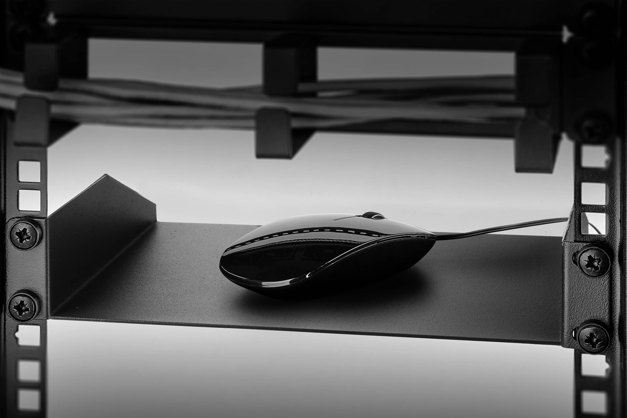Een zwarte computermuis ligt op een metalen plank onder een tafel met kabels op de achtergrond.
