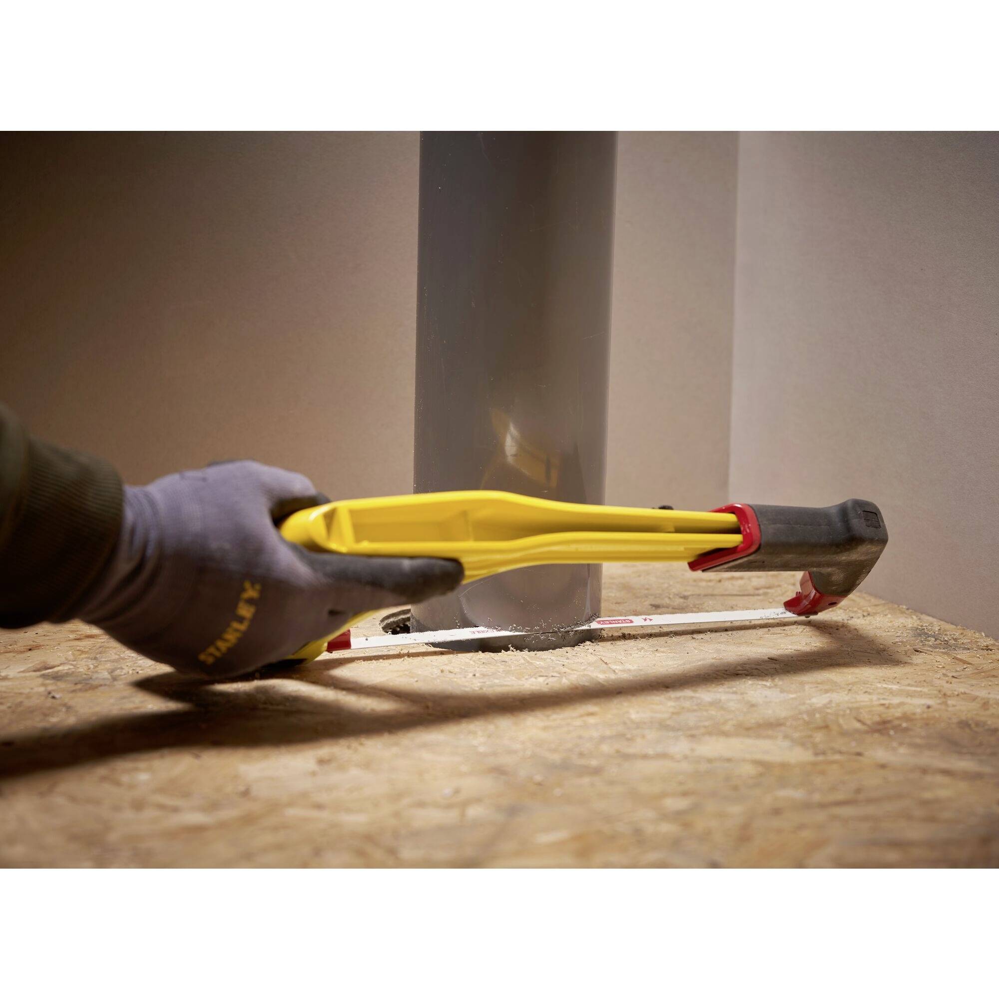 Een persoon draagt werkhandschoenen en gebruikt een geel en rood gereedschap om iets met een hamer vast te maken op een houten oppervlak.