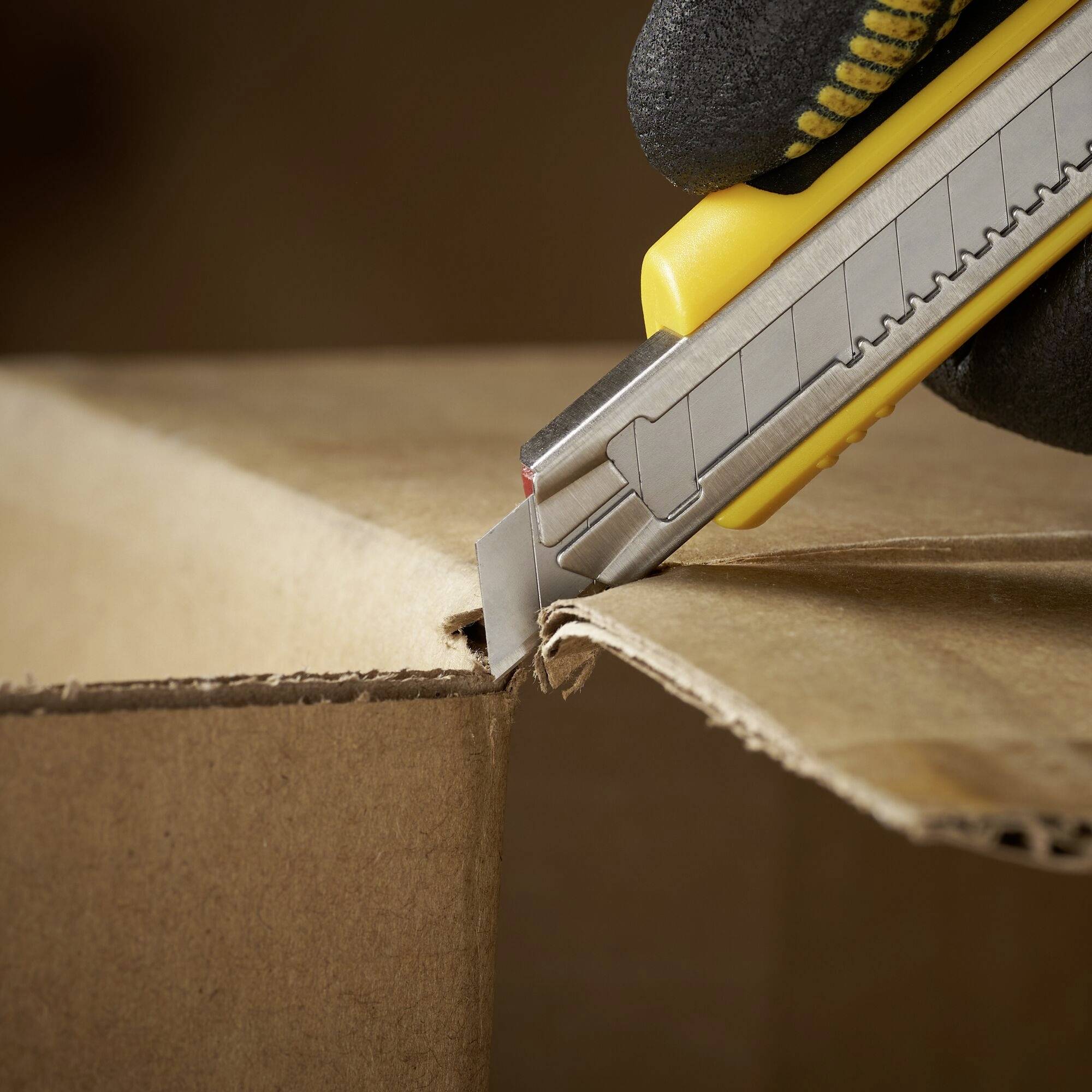 Een hand met een geel stanleymes snijdt voorzichtig een kartonnen doos langs de rand open.