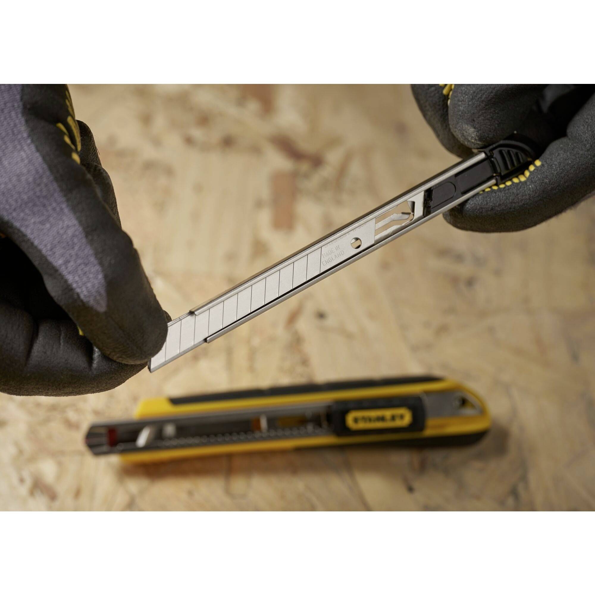 Een handsnijder met meerdere messen wordt vastgehouden door iemand met beschermende handschoenen. Een gele cuttermes ligt op de achtergrond.