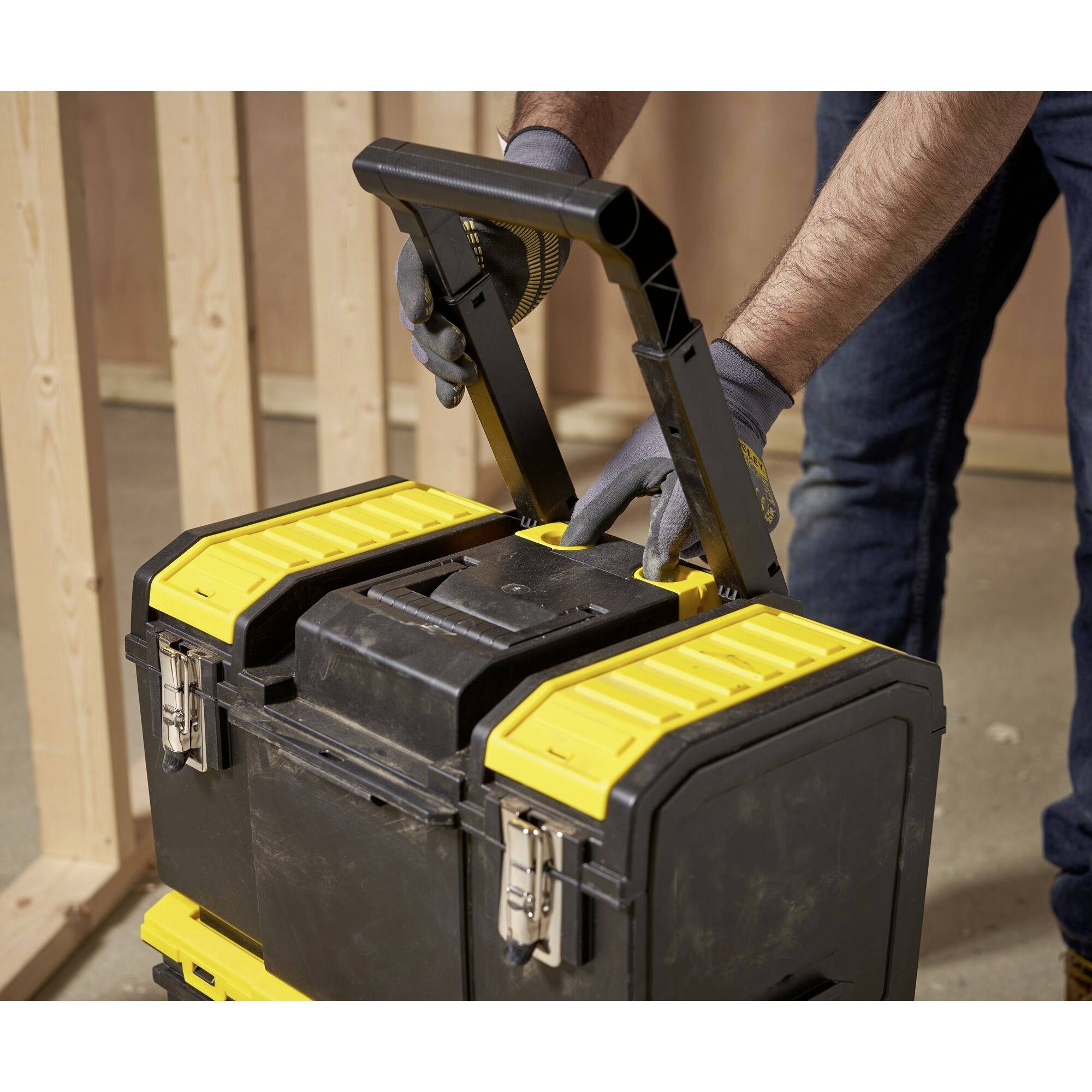 Een persoon met handschoenen tilt een geel-zwarte gereedschapskist op aan een handgreep. Op de achtergrond is een houten frame zichtbaar.