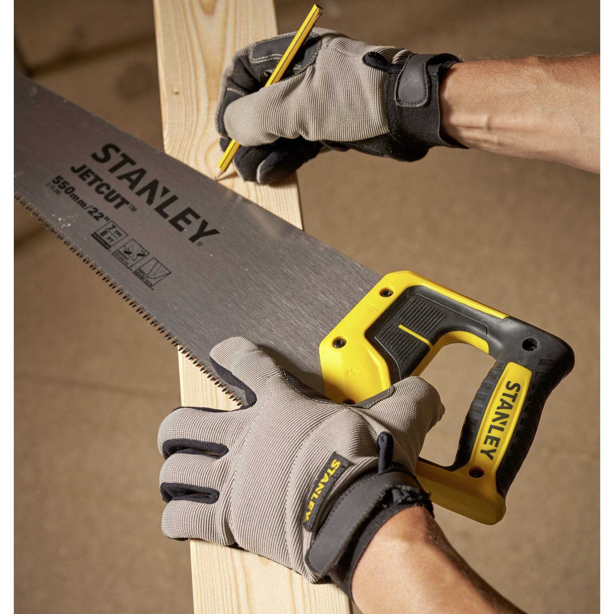 Een persoon met werkhandschoenen markeert een houten plank met een potlood, terwijl hij een handzaag met een gele handgreep vasthoudt.