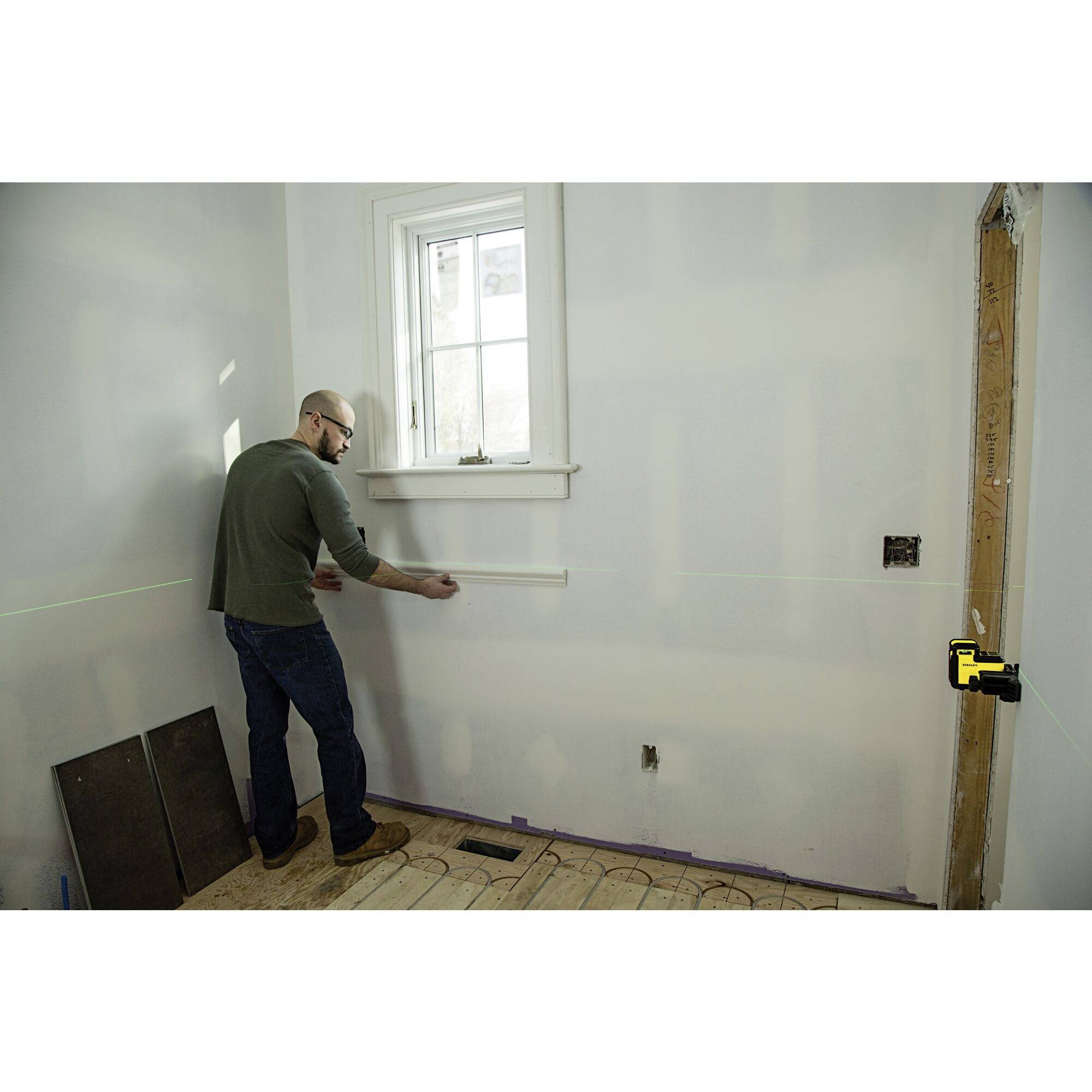 Een man meet een muur met een meetlint in een gerenoveerde ruimte. Op de muur zijn laserlijnen aangebracht voor nauwkeurige uitlijning.