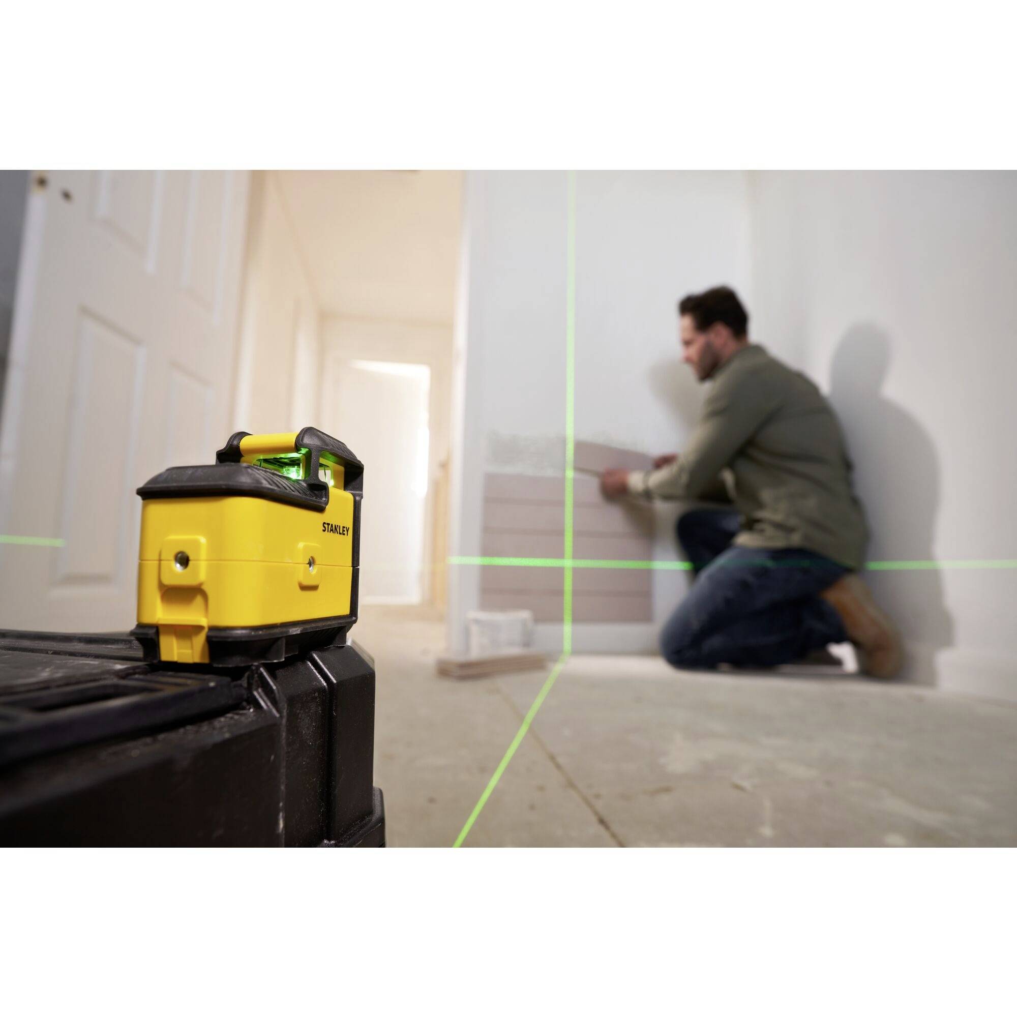 Een man gebruikt een laserwaterpas om tegels aan een muur te plaatsen. Het apparaat projecteert groene lijnen voor nauwkeurige uitlijning.