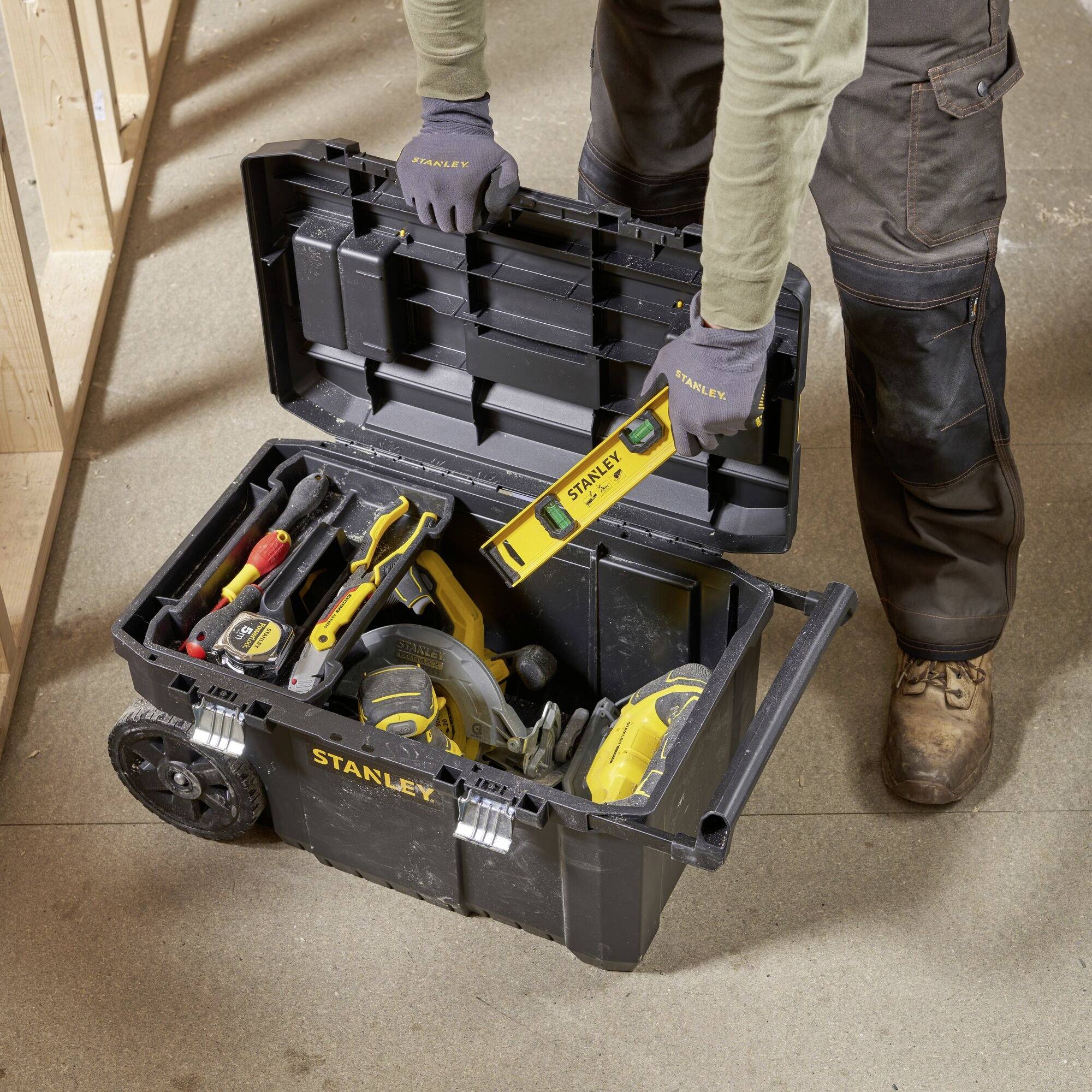 Een persoon opent een gereedschapskoffer op wielen. Daarin bevinden zich verschillende gereedschappen, waaronder een handhefelzaag.