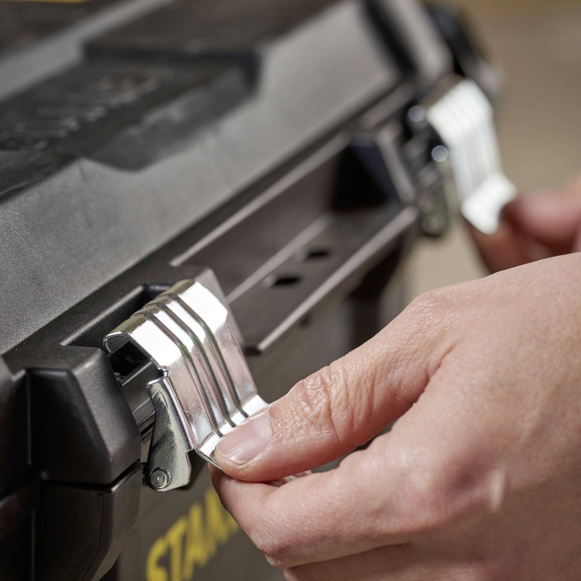 Close-up van een hand die de sluiting van een zwarte gereedschapskist opent. De kist heeft zilveren metalen sluitingen.