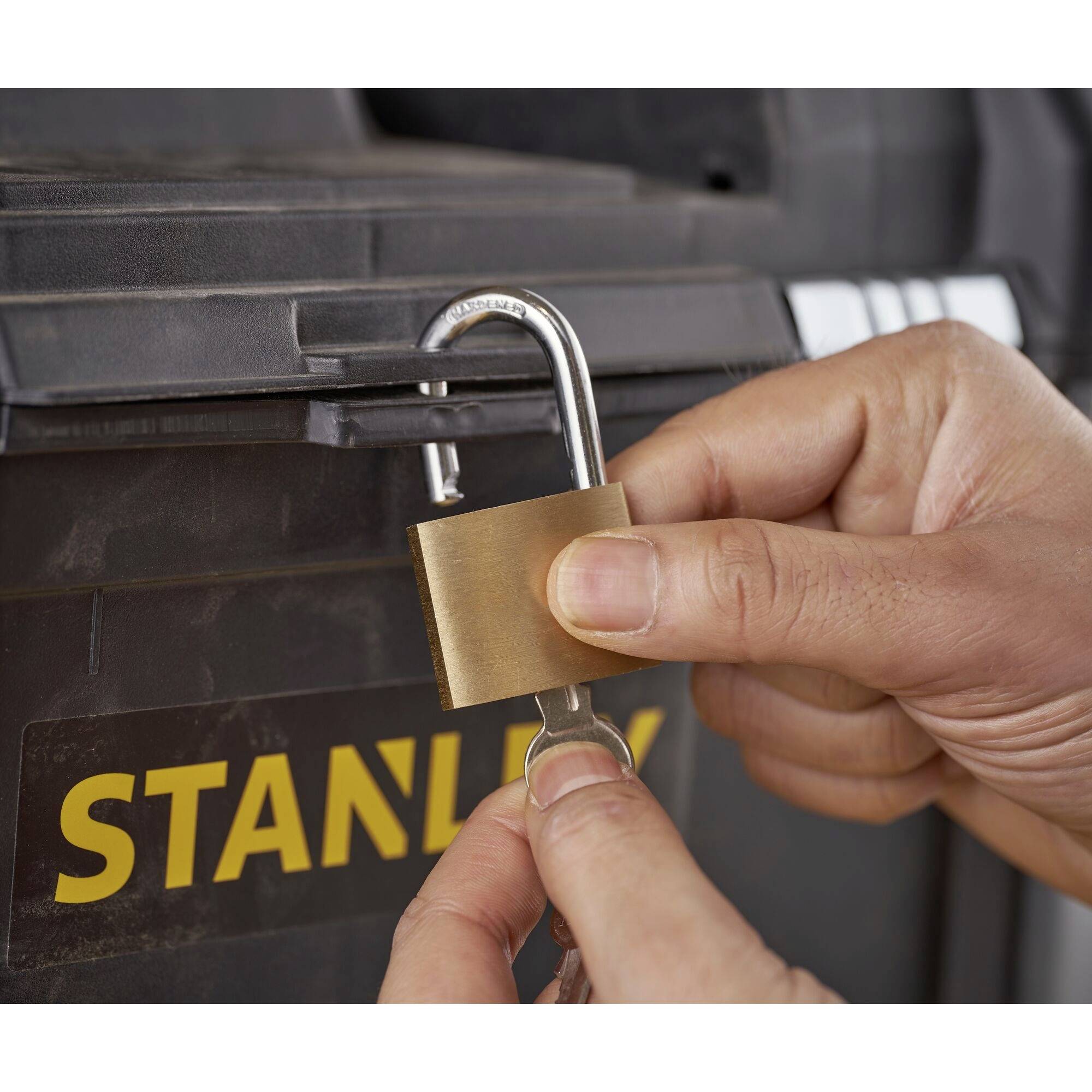 Twee handen sluiten een hangslot aan op een zwarte kist met het opschrift 'STANLEY'.