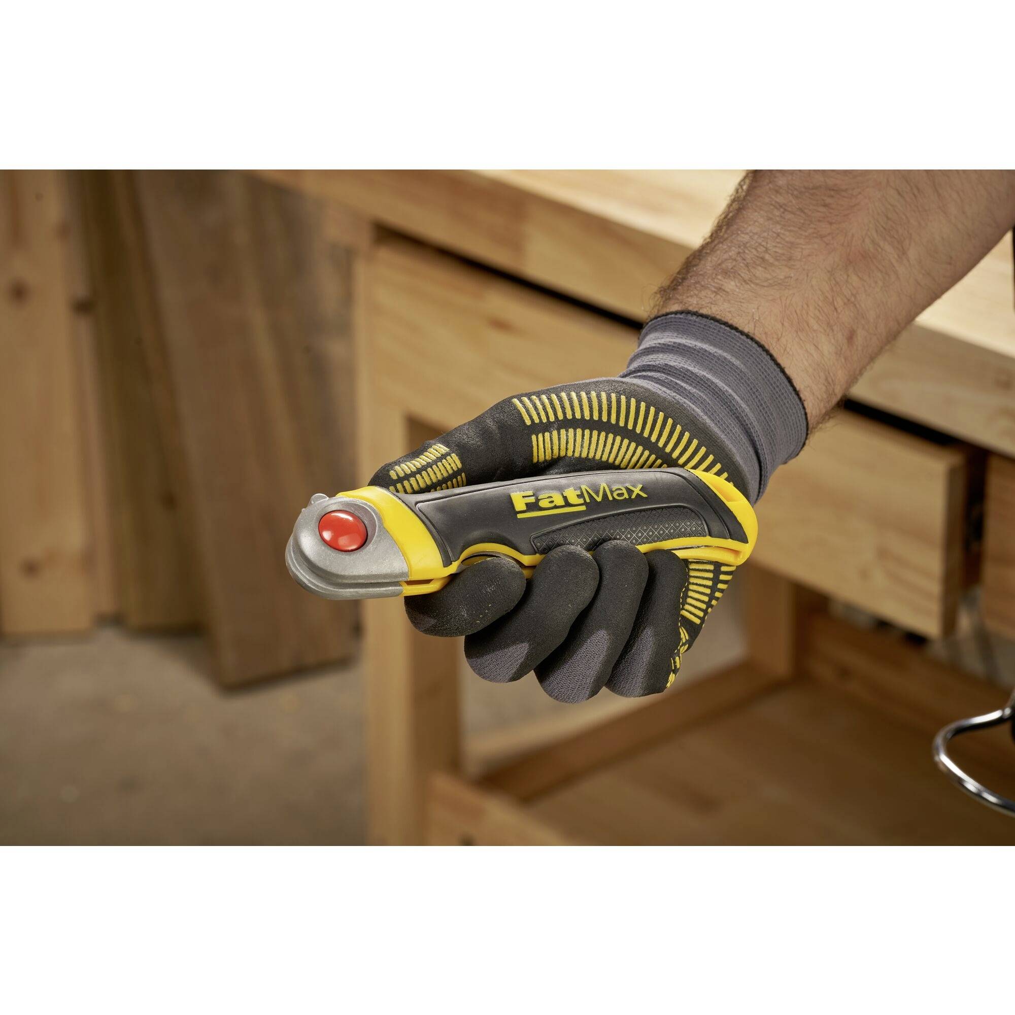 Een hand draagt een geel en zwarte handschoen en houdt een geel FatMax veiligheidsmesmes met een rode knop voor een werktafel.