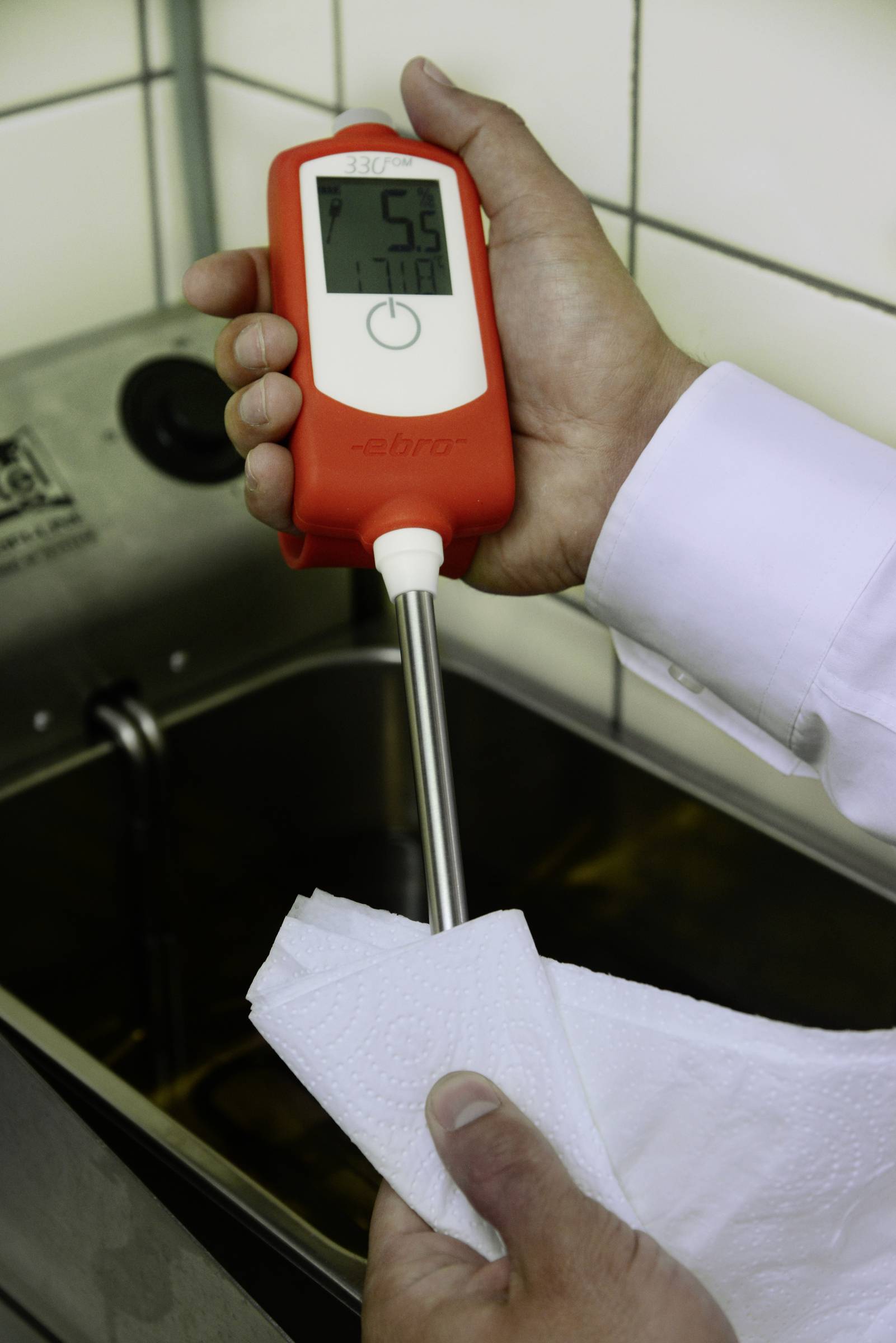 Een persoon houdt een digitale thermometer vast die 56°C aanwijst, naast een bekken met water; de metalen sonde wordt schoongeveegd.