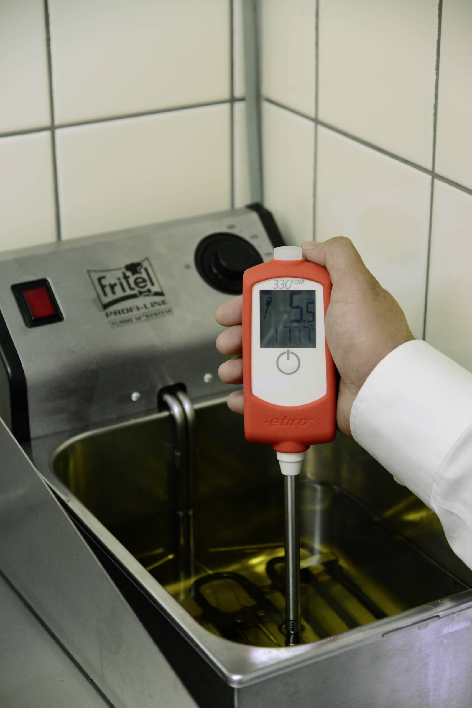 Een persoon houdt een thermometer boven een friteuse en meet de temperatuur van de olie. Het display toont 75,6 graden aan.