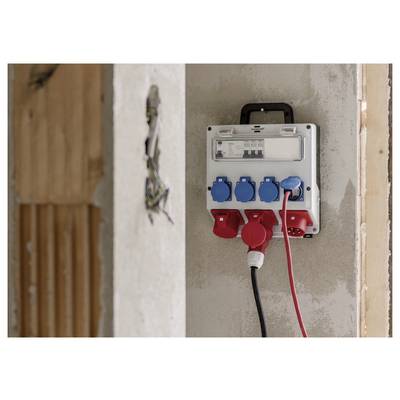 Aan de muur gemonteerde elektrische stopcontactdoos met blauwe en rode contactdozen in een gedeeltelijk afgewerkte binnenmuur, met ingesteken snoeren.