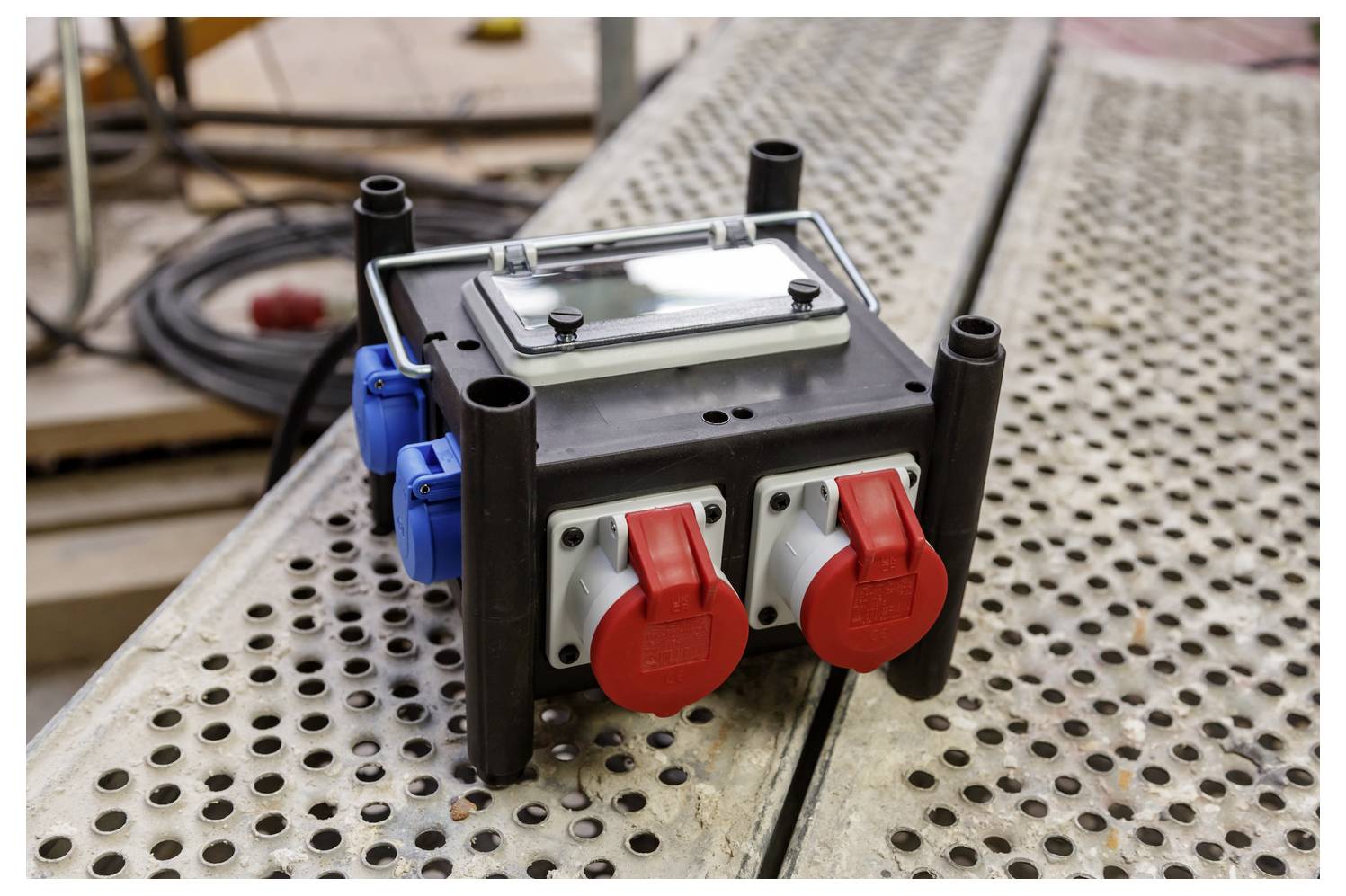Draagbare elektrische verdeelkast met rode en blauwe stekkercontacten op een geperforeerd metalen werkoppervlak.