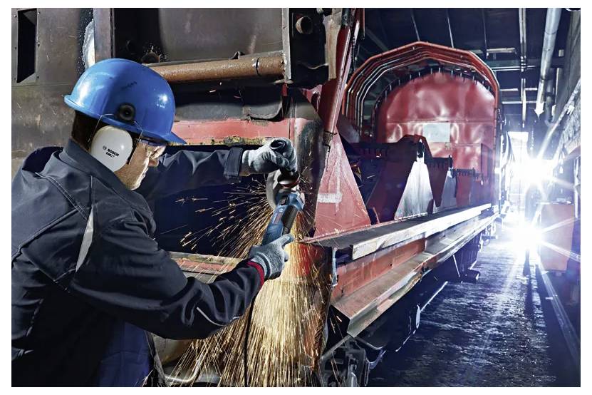 Een werknemer in beschermende uitrusting slijpt metaal in een industriële omgeving, met felle vonken die rondvliegen. De omgeving oogt groot en volgestouwd met machines.