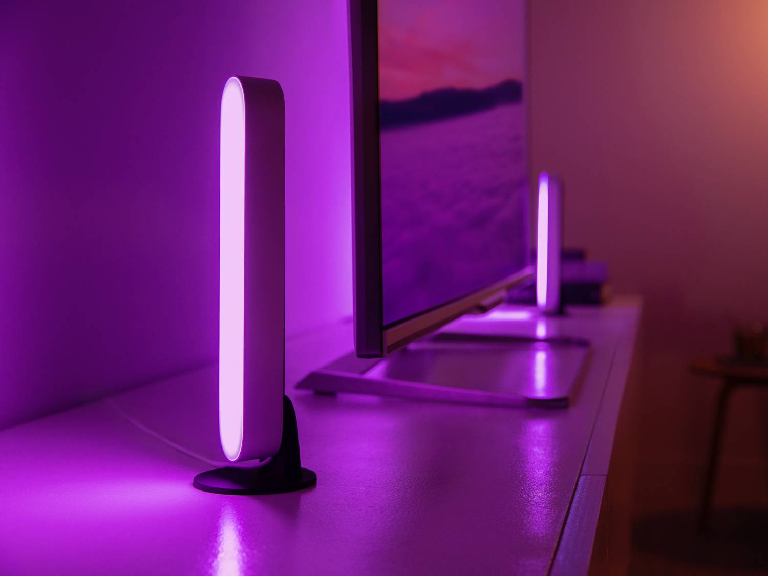 Twee slanke, verticaal geplaatste LED-lampen schijnen in intens paars naast een beeldscherm op een modern bureau.