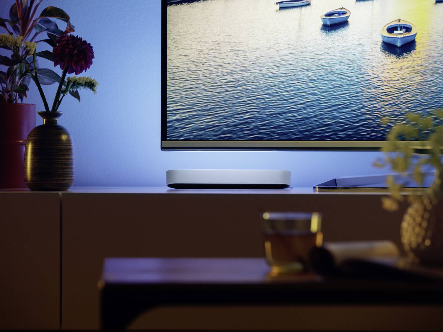Een woonkamer met een televisie op een rek, daaronder een soundbar. Op de tafel op de voorgrond staat een drankje. Op de achtergrond toont het scherm een afbeelding van boten op het water.