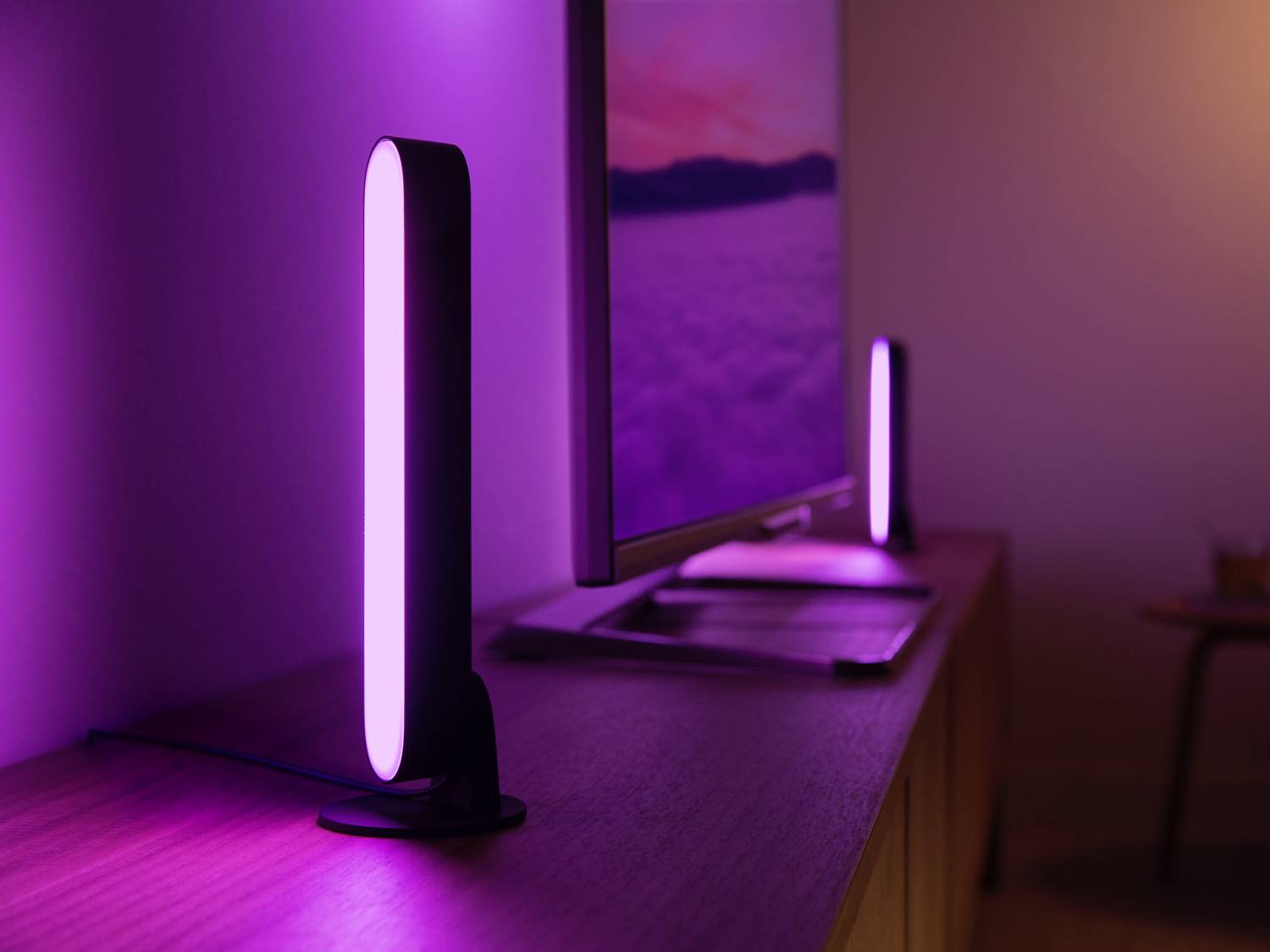 Twee verticale, lichtgevende LED-lampen op een tafel naast een scherm dat een kleurrijk landschapstafereel vertoont.
