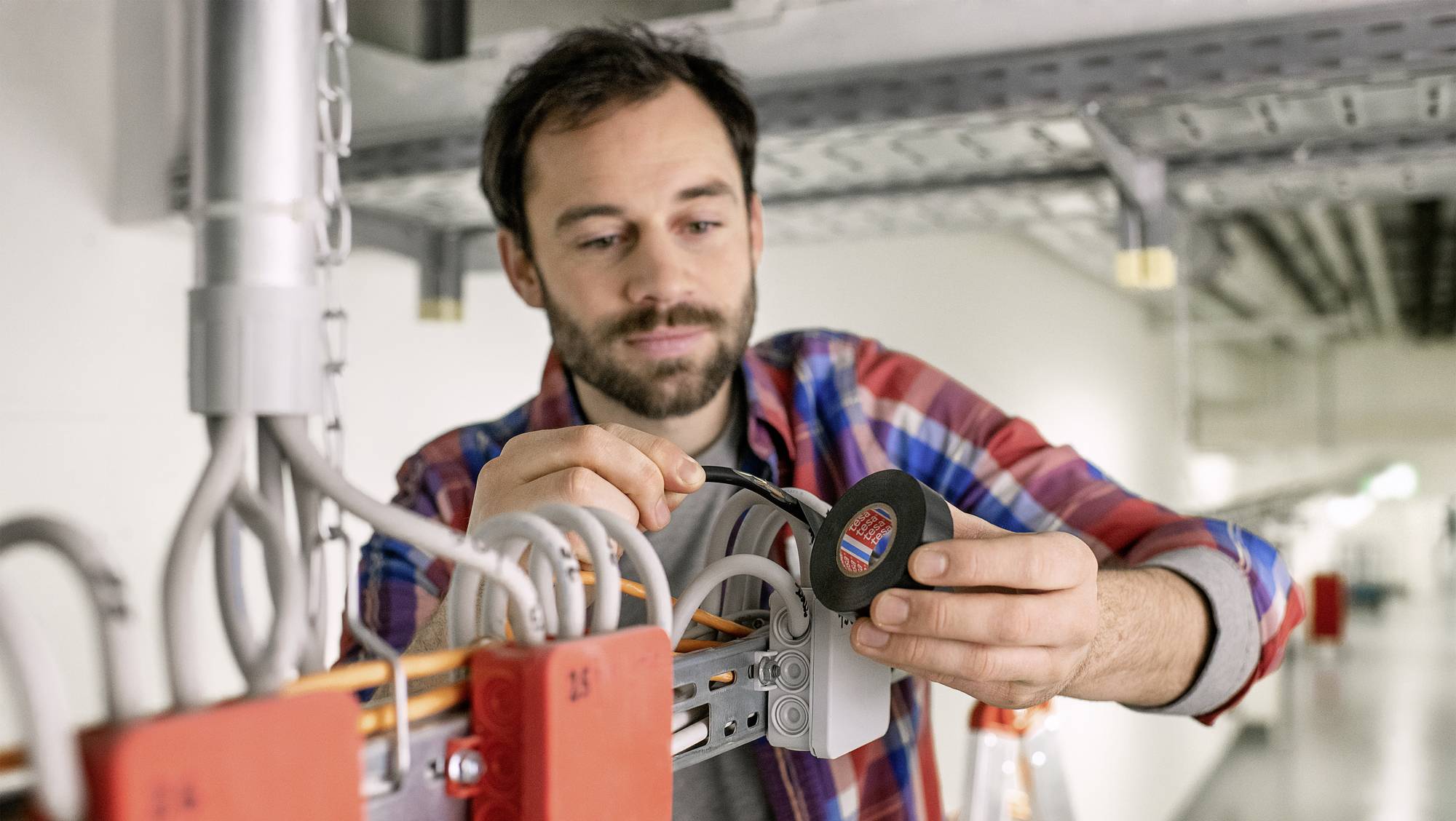 Een man controleert elektrische kabels in een gebouw. Hij draagt een overhemd en houdt een meetapparaat in zijn hand.