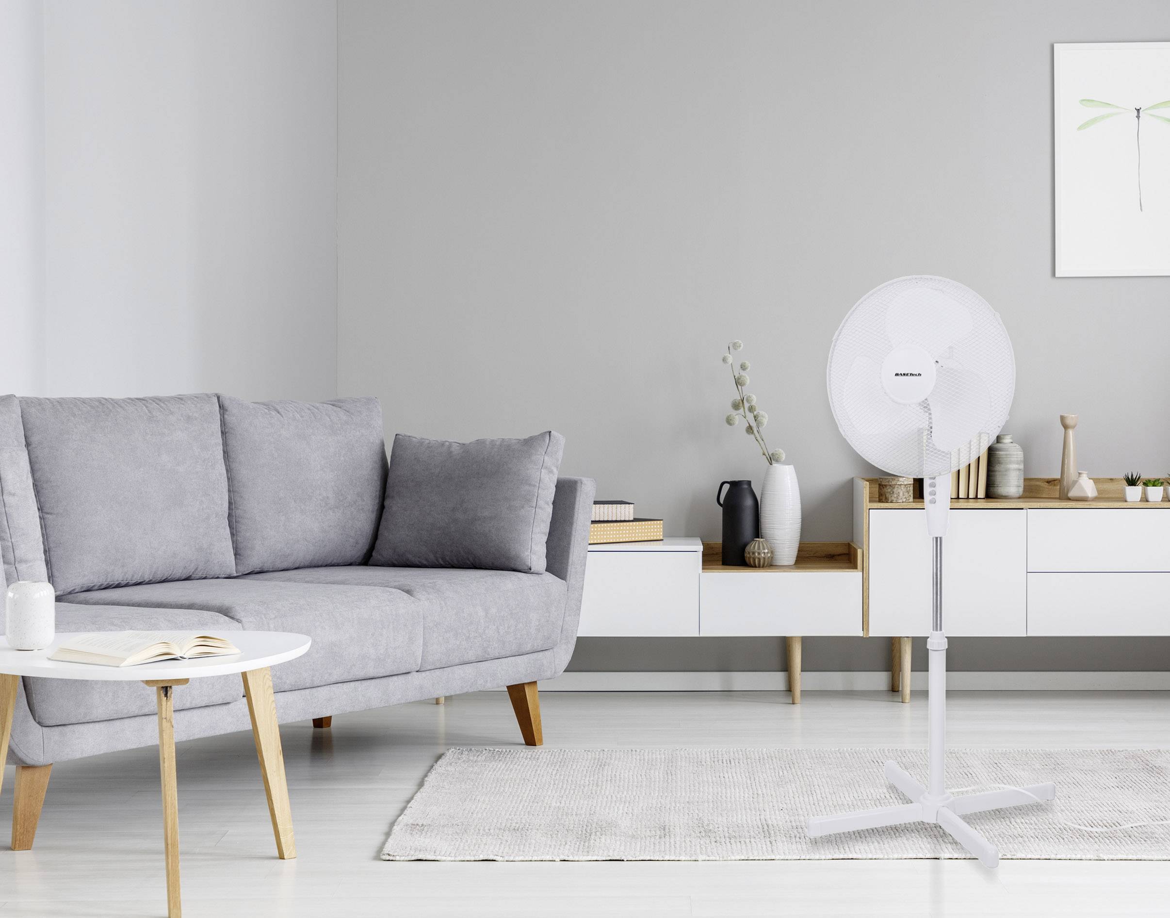 Grijze woonkamer met bank, ronde tafel, vloerkleed en staande ventilator. Minimalistische stijl met sobere decoratie, lichte muren.