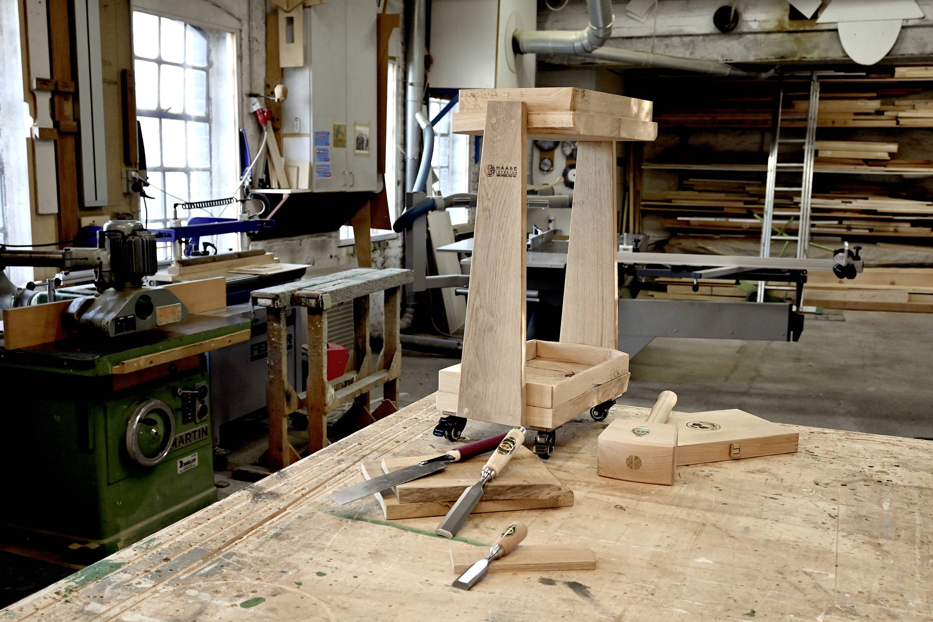 Een houten werkplaats met een onafgewerkte houten kruk op een werkbank, omringd door gereedschappen en machines voor houtbewerking.
