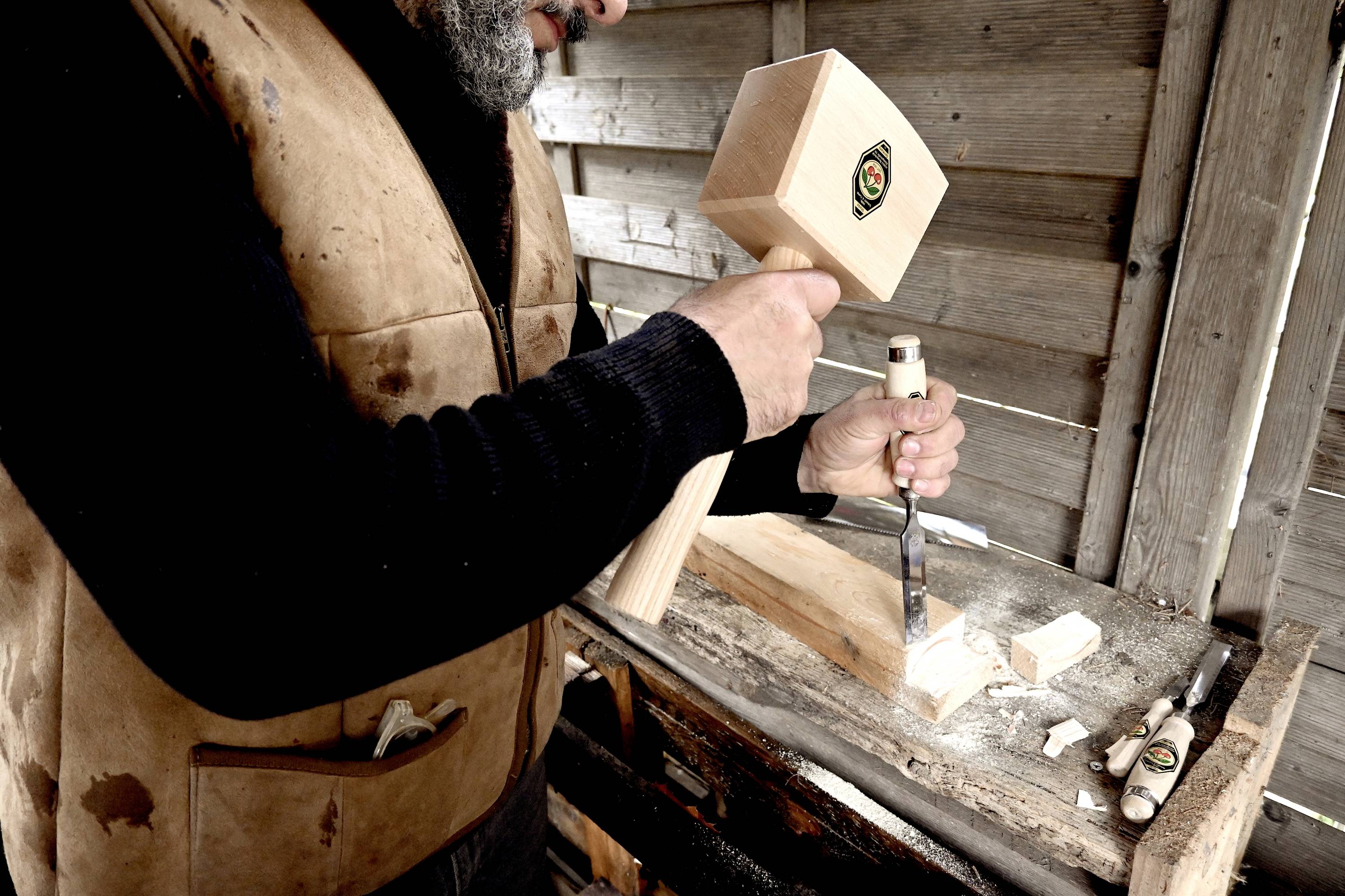 Een man bewerkt een stuk hout met een beitel en een houten hamer in een werkplaats. Hij draagt een bruine vest.