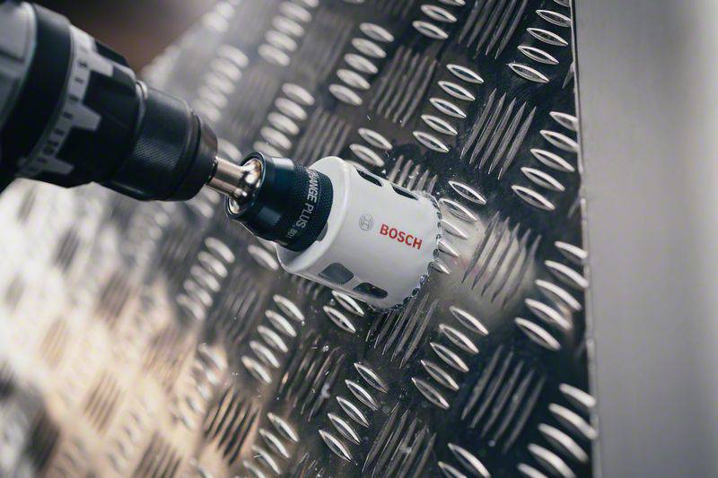 Boormachine met gatenzaag boort in metalen plaat met geprofileerd plaatpatroon. Merknaam 'Bosch' zichtbaar op de gatenzaag.