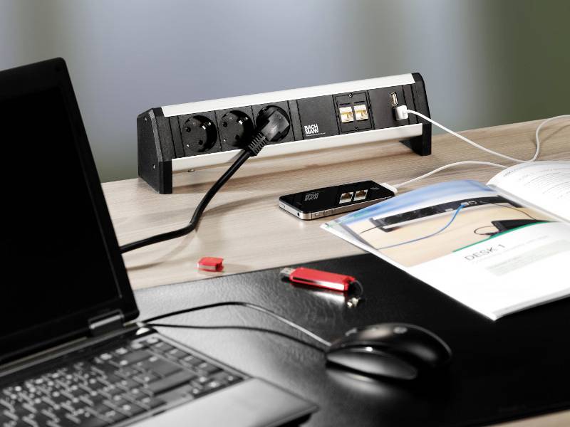 Laptop en muis op een bureau naast een stekkerdoos met twee stopcontacten en een USB-aansluiting. Mobiele telefoon wordt opgeladen.