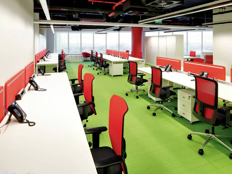 Een modern, leeg kantoor met rode en groene kleuraccenten. Rijen bureaus met telefoons en bureaustoelen, grote ramen op de achtergrond.