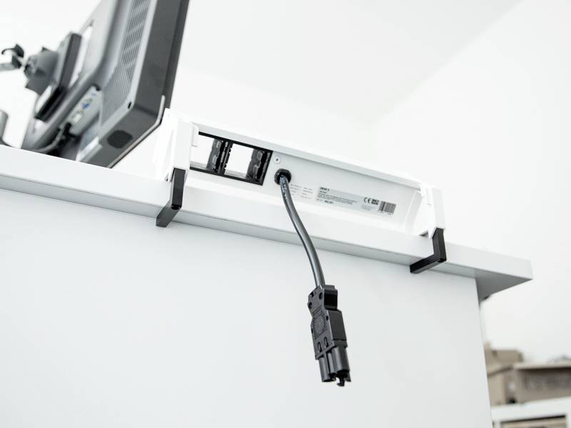 Een stroomkabel is aangesloten op een stopcontact aan de onderkant van een bureau met een computermonitor bovenop, wat wijst op een kantooropstelling.