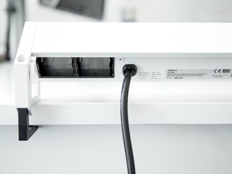 Een kabel is aangesloten op een wit apparaat met twee rechthoekige sleuven. Naast de kabel is een etiket met tekst en symbolen zichtbaar.