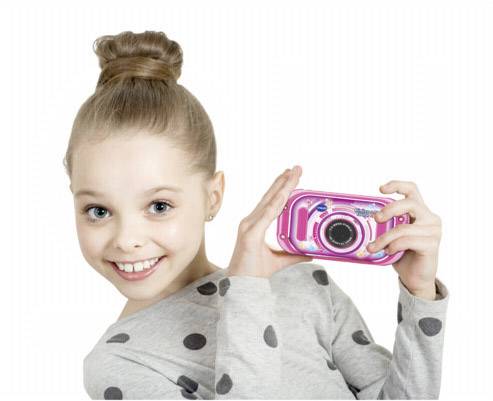 Een meisje met haar in een hoge knot glimlacht naar de camera en houdt een roze speelgoedcamera vast.
