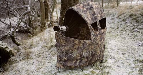 Een gecamoufleerd tent met een uitgeklapt objectief staat in het besneeuwde bos, klaar voor natuurfotografie.