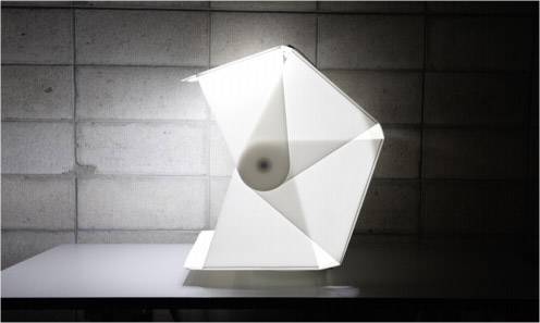 Een modern design-lichtobject op een tafel voor een betonnen muur, gevormd uit gevouwen geometrische vormen die een warm licht uitstralen.