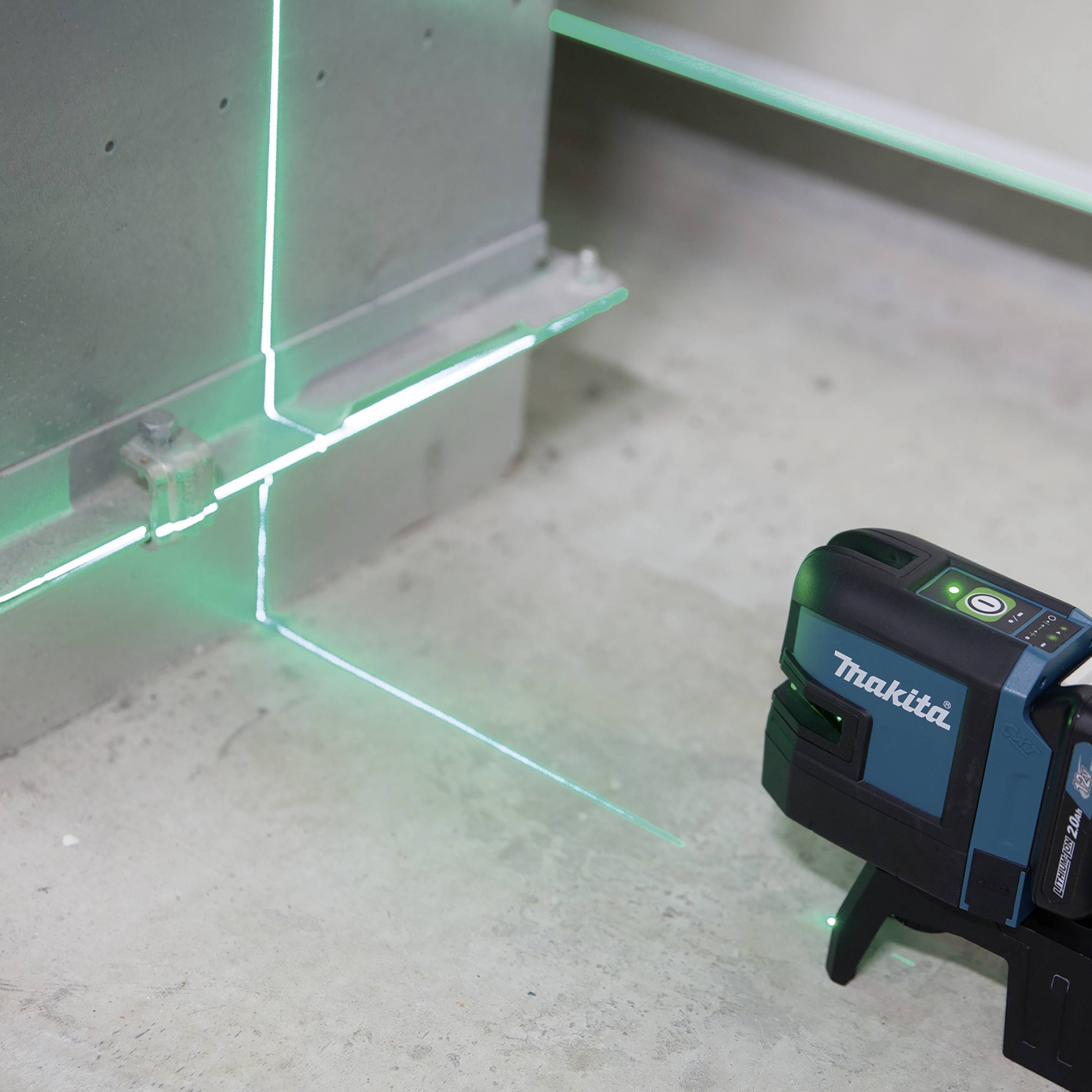 Een groene laser waterpast op een betonnen vloer, projecteert lijnen op de muur en de vloer. Een zwart laserapparaat is zichtbaar.