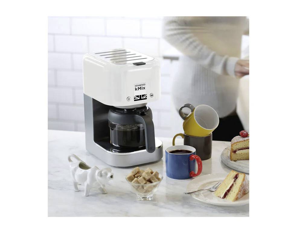 Koffiemachine op keukentafel met gevulde kan, een kopje koffie, suikerklontjes en een aangesneden taart op een bord.