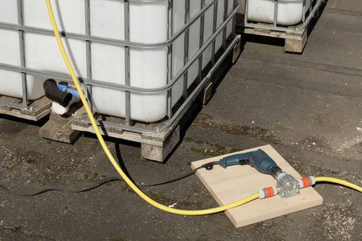 Een pomp pompt vloeistof door een gele slang uit een grote watertank. De pomp staat op een houten plank.