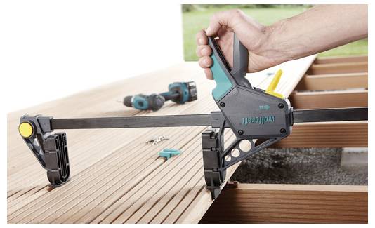 'Een hand die een Wolfcraft klem gebruikt om een houten plank vast te zetten. Gereedschappen en schroeven zijn zichtbaar op de achtergrond, wat wijst op een constructieopstelling.'