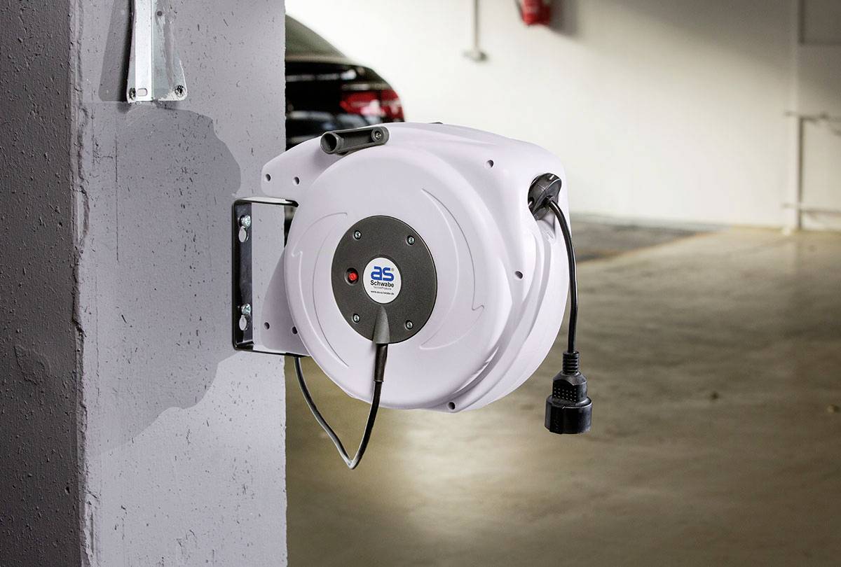 Wandgemonteerde kabelrol in een parkeergarage, bevestigd aan een betonnen zuil; een zwarte auto is op de achtergrond zichtbaar.