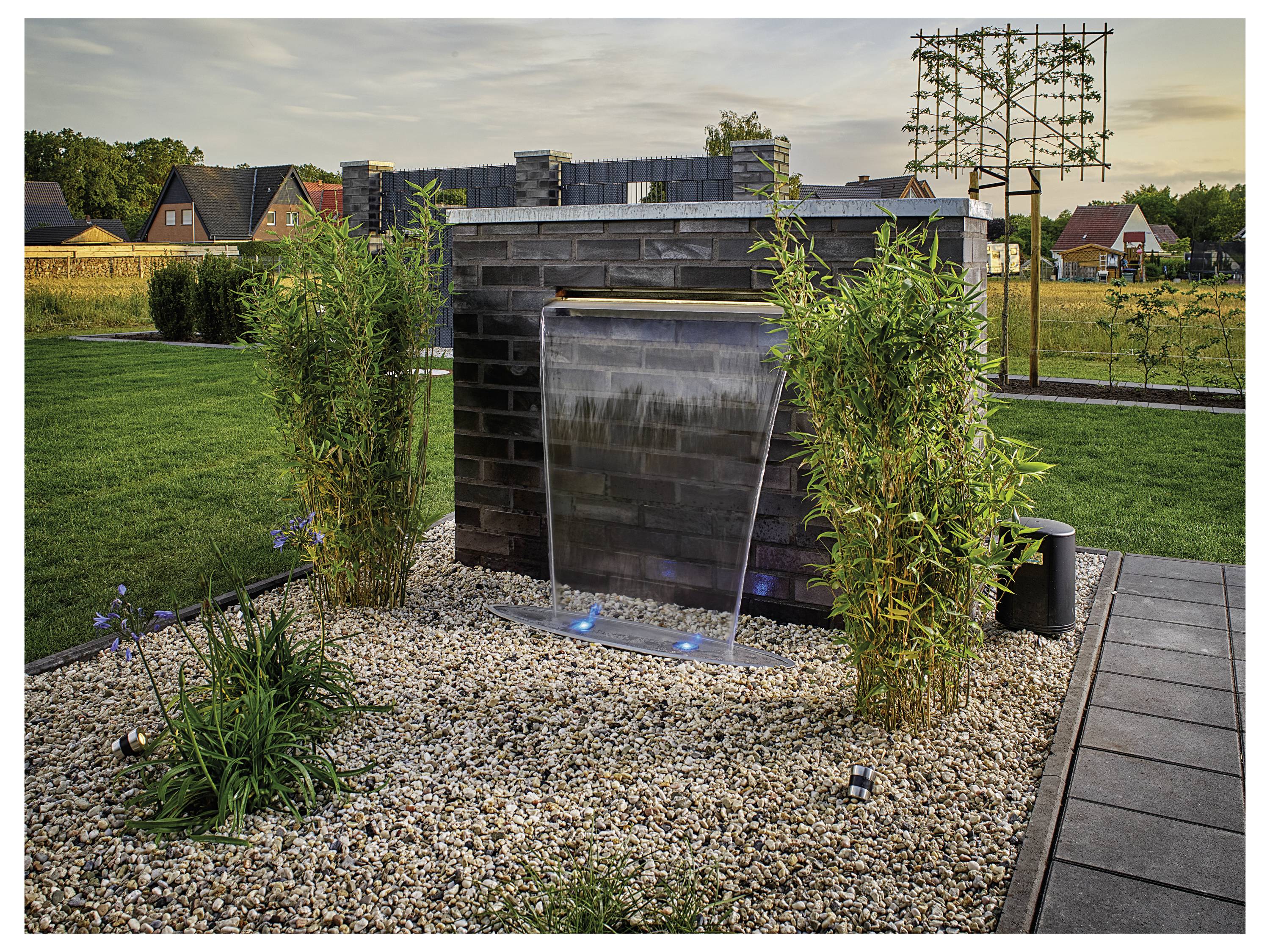 'Een kleine tuinwaterpartij met een waterval die trapsgewijs naar beneden valt, omgeven door groene planten en kiezels. Huizen en bomen zijn zichtbaar op de achtergrond.'
